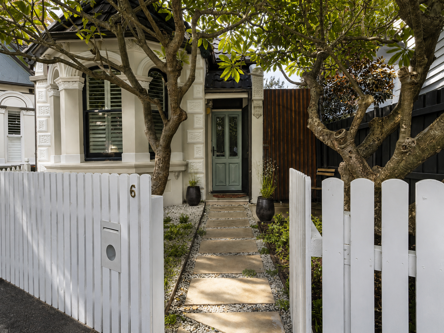 Limestone path through white picket gate to heritage cottage with mature trees and low plantings